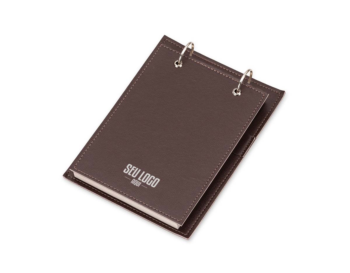 Bloco de anotações de mesa com trilho de ferro e capa em couro sintético. Contém suporte interno lateral para caneta (não inclusa) e cerca de 96 folhas recicladas com área destacável para facilitar o uso no dia a dia.